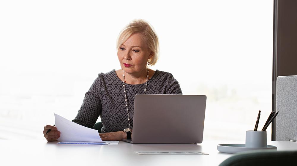  woman at the computer is reading a paper document.