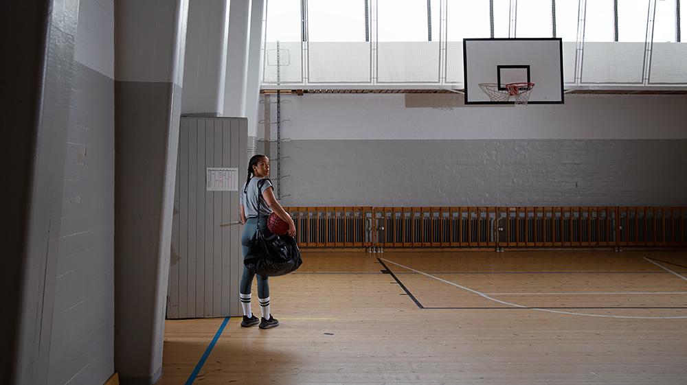 Nuori tyttö yksin seisoo tyhjässä urheilusalissa koripallo kainalossaan.